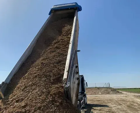 La plataforma de un camión volquete se levanta y descarga una pila de astillas de madera contra un cielo azul.