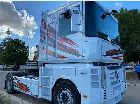 Camión semirremolque blanco con calcomanías rojas y naranjas estacionado al costado de una carretera en un día soleado.