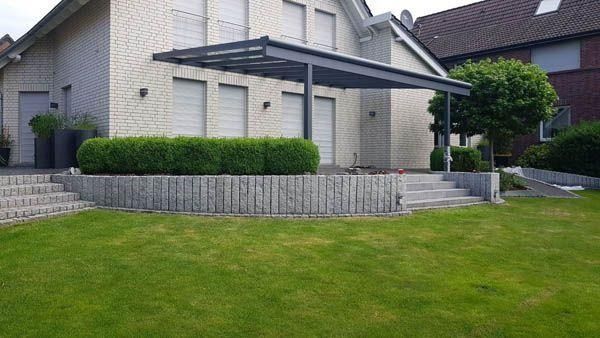 Ein modernes Haus mit überdachter Terrasse, Garten und Stützmauer aus Betonsteinen.