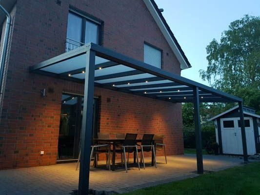 Terrasse mit dunkelgrauer Pergola, Gartentisch, Stühlen und Beleuchtung vor einem Backsteinhaus.