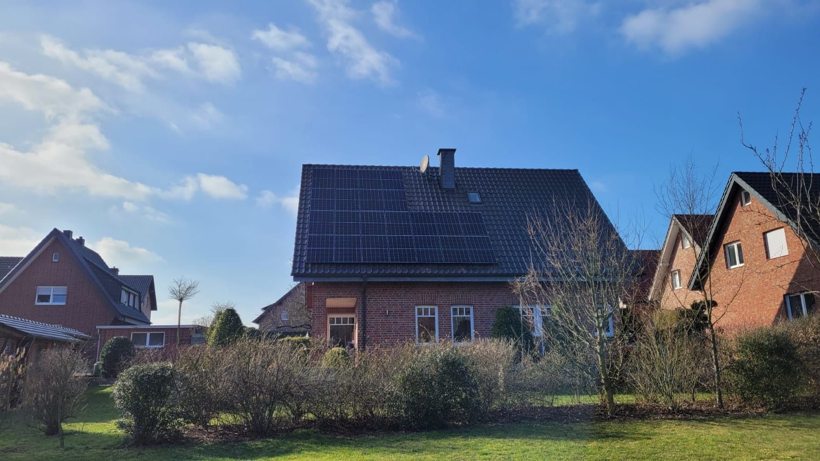 Backsteinhaus mit Solarpaneelen auf dem Dach, unter blauem Himmel, davor von Büschen umgeben.
