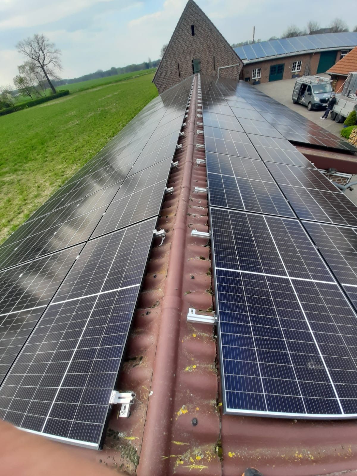 Auf einem roten Metalldach, neben einem braunen Dachfirst, sind Solarpaneele installiert. Im Hintergrund sind eine grüne Wiese und ein Gebäude zu sehen.