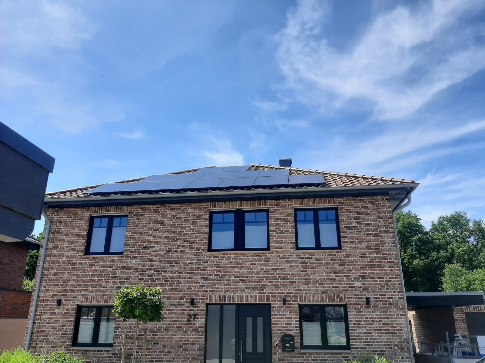 Zweistöckiges Backsteinhaus mit Solarpaneelen auf dem Dach unter blauem Himmel. Schwarze Fensterrahmen und Tür.