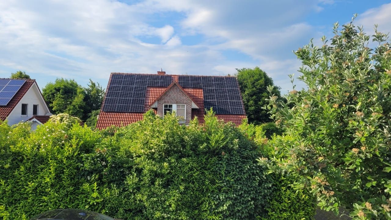 Unter einem bewölkten Himmel liegt hinter grünen Büschen ein Haus mit Ziegeldach.