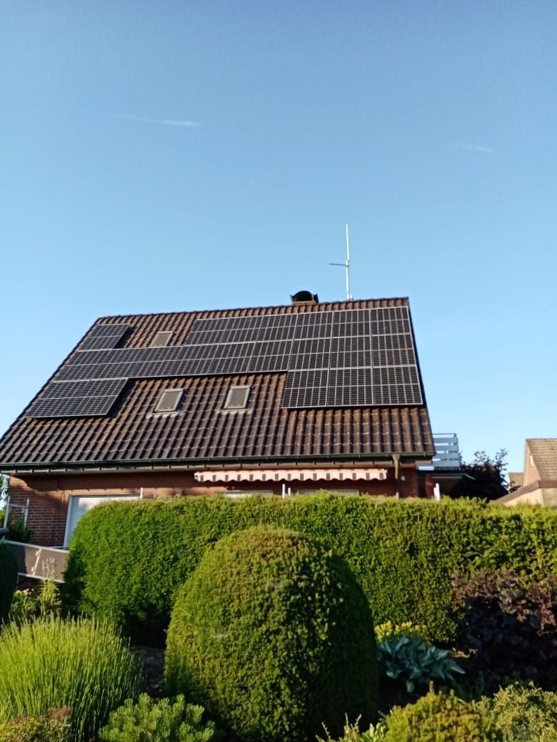 Haus mit Solarpaneelen auf dem Dach, blauem Himmel und grüner Hecke.