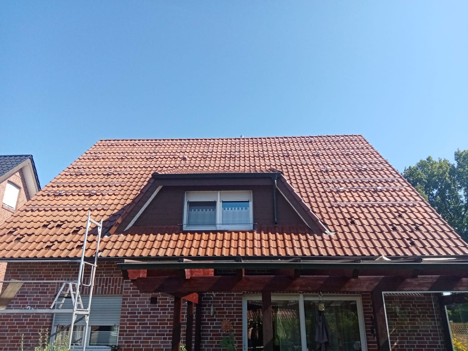 Das rotziegelgedeckte Dach eines Backsteinhauses mit Dachgaube und Pergola; darüber ein klarer blauer Himmel.
