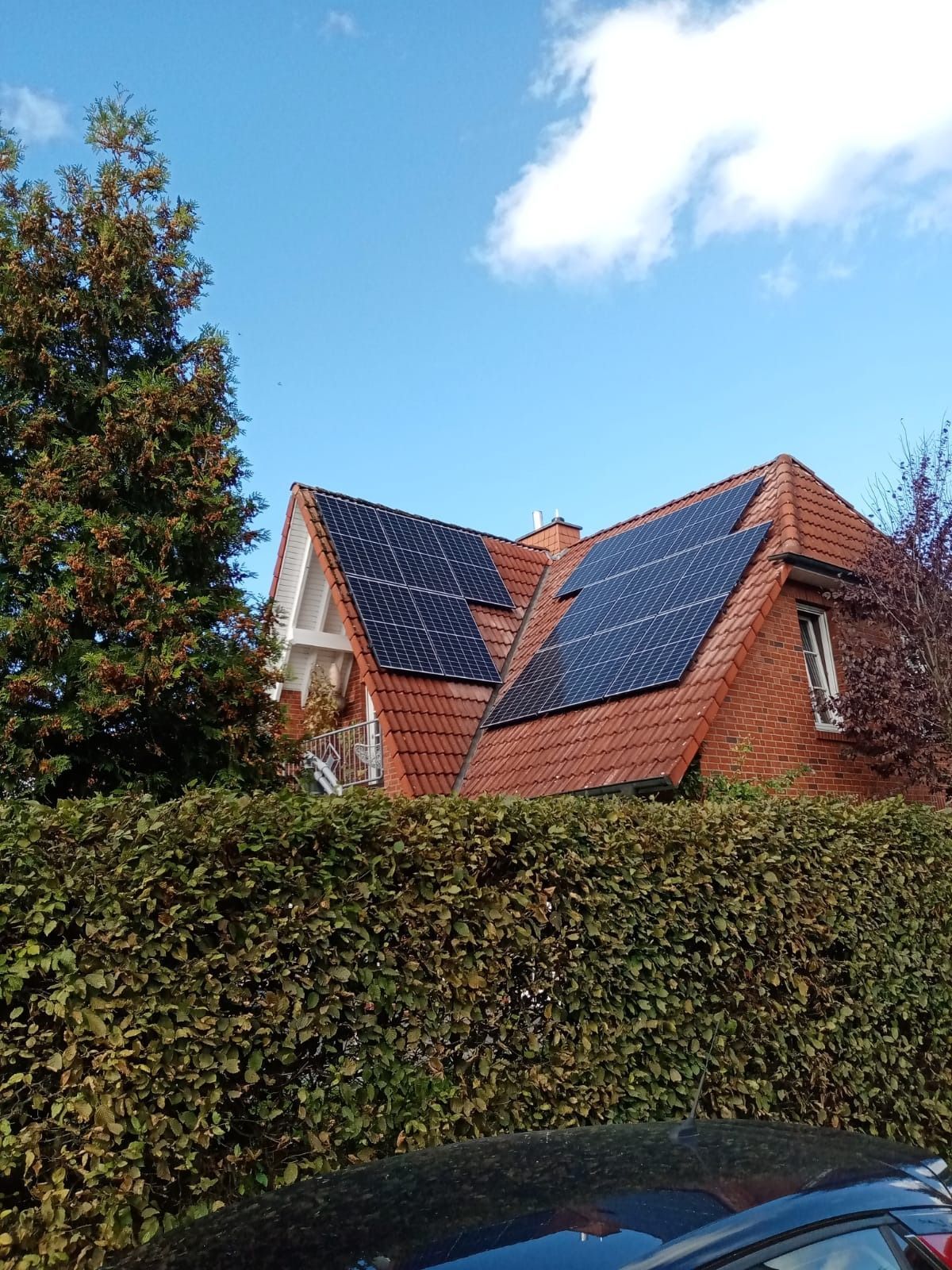 Solarpaneele auf einem roten Ziegeldach vor strahlend blauem Himmel. Grüne Hecke und Bäume im Vordergrund.