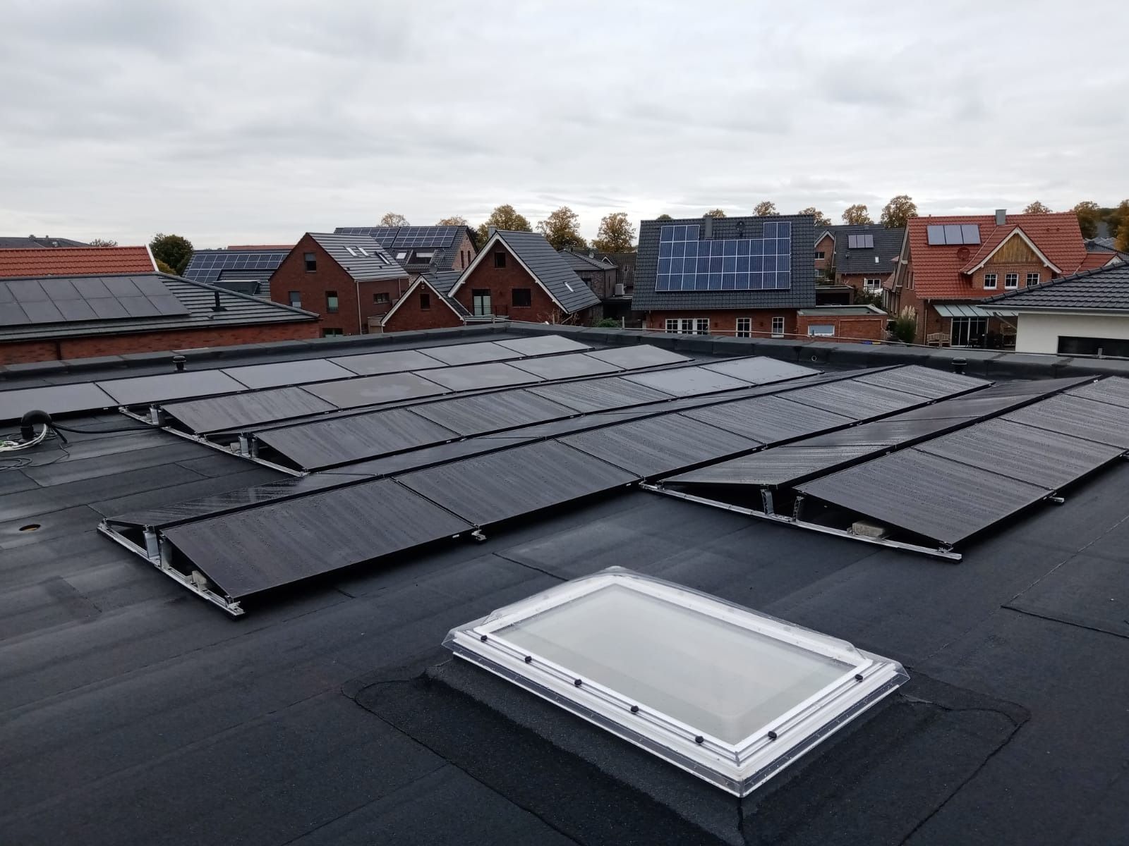 Auf einem Flachdach mit Oberlicht sind Solarpaneele installiert; im Hintergrund sind weitere Gebäude und ein bewölkter Himmel zu sehen.