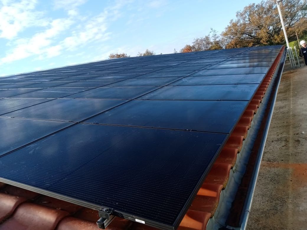 Auf einem Ziegeldach sind Solarpaneele installiert. Die Paneele sind dunkel, die Ziegel orangebraun. Darüber strahlend blauer Himmel.