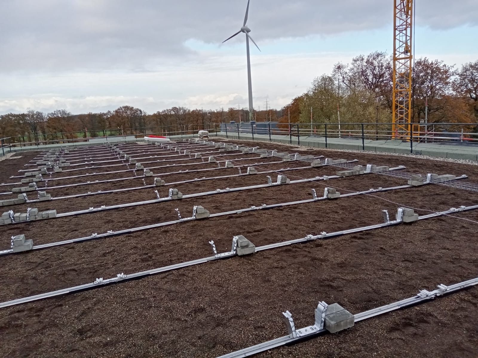 Dach mit für die Installation von Solarmodulen vorbereiteter Metallkonstruktion, im Hintergrund eine Windkraftanlage und ein Kran.