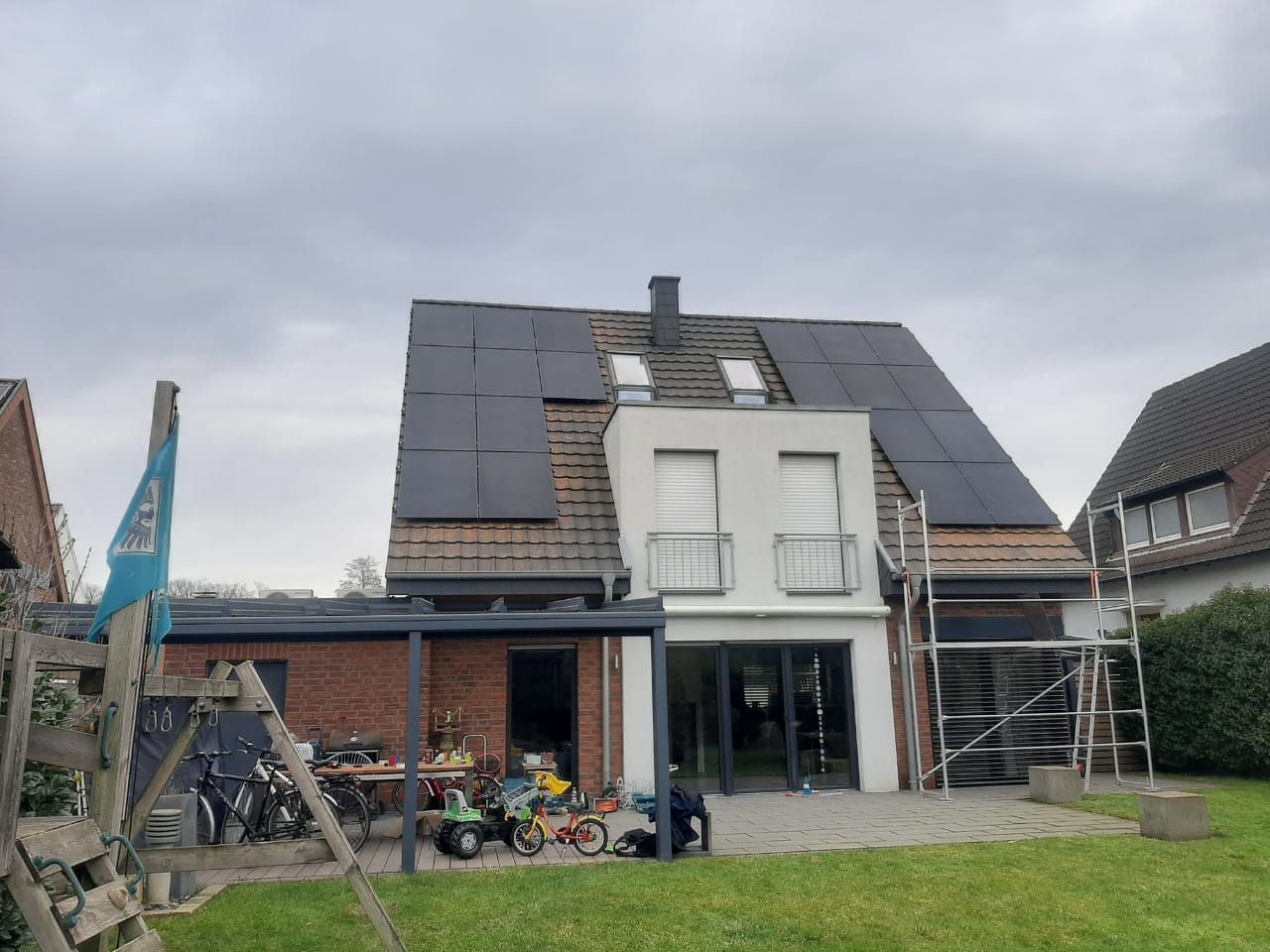 Haus mit Solarpaneelen auf dem Dach, Gerüst und einem kleinen Hof mit Spielzeug und einer Pergola. Bewölkter Himmel.