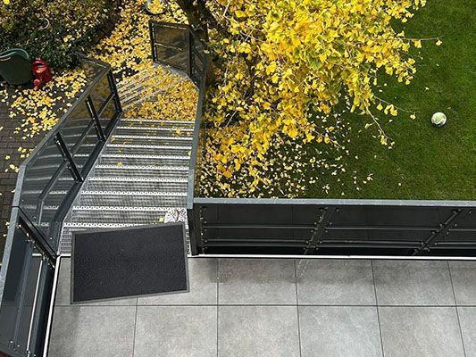 Draufsicht auf eine Außentreppe, die zu einer grünen Rasenfläche mit einem Baum und verstreuten gelben Blättern führt.