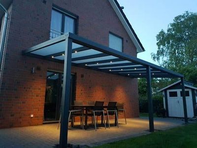 Terrasse mit Pergola, Tisch und Stühlen. Außenansicht des Backsteinhauses mit Deckenbeleuchtung in der Dämmerung.