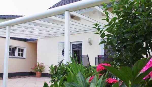 Weiße Terrassenmarkise über einer Terrassentür und Fenstern, im Vordergrund ein beigefarbenes Gebäude, rosa Blumen und Grünpflanzen.