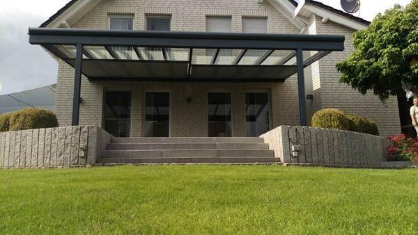 Hinterhof mit überdachter Terrasse, Treppe und Haus. Grüner Rasen und Sträucher.