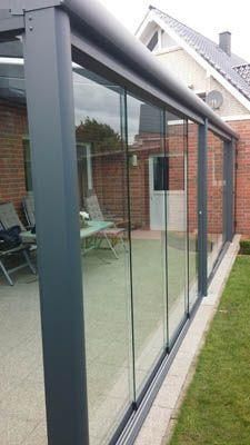 Verglaste Terrassenüberdachung mit grauem Rahmen vor einem Backsteingebäude, Blick auf Terrassenmöbel und Rasen.