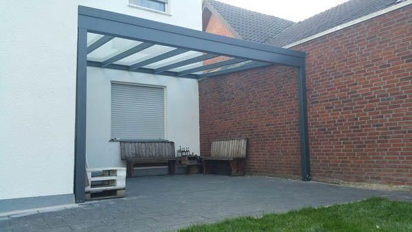 Eine graue Pergola überspannt eine Terrasse mit Ziegel- und weißen Wänden, zwei Holzbänken und einem Betonboden.