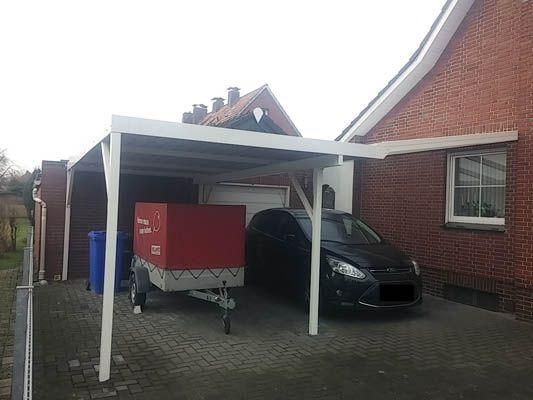 Carport an einem Backsteinhaus, darunter ein Auto geparkt, links davon ein roter Anhänger.