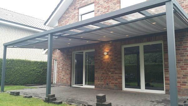 Graue Pergola über einer Ziegelterrasse mit Glastüren, eingebauter Beleuchtung und grüner Hecke.