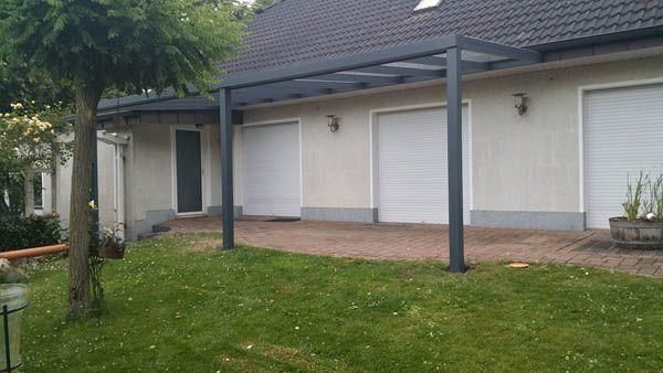 Graue Pergola über einer Ziegelterrasse vor einem Haus mit geschlossenen weißen Garagentoren. Grüner Rasen.