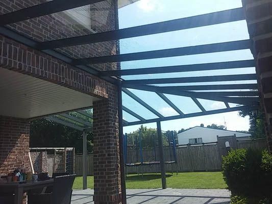 Überdachte Terrasse mit transparentem Dach, Backsteinsäulen und Blick auf einen Hinterhof mit Trampolin.