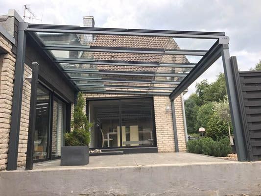 Terrasse mit Glasdach über Tür und Fenstern an einem Backsteinhaus. Graue Stützen und dunkle Zierleisten.