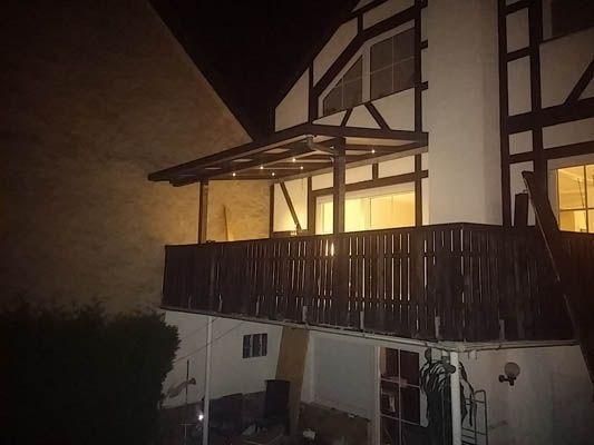 Balkon eines Hauses bei Nacht, Holzgeländer und Pergola, drinnen Licht an.