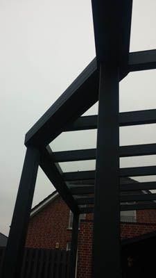 Schwarze Pergola vor bewölktem Himmel, im Hintergrund ist ein Backsteingebäude zu erkennen.