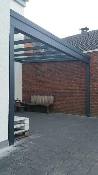 Eine graue Pergola mit Glasdach ist an einer Backsteinmauer befestigt. Darunter steht eine Holzbank.