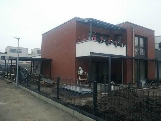 Modernes zweistöckiges Haus mit Balkon und dunklem Metallzaun davor. Die Fassade ist rotbraun. Bewölkter Tag.