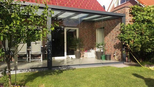 Terrasse mit Glasdach und Schiebetüren, Backsteinmauer, grüner Rasen und Bäume.