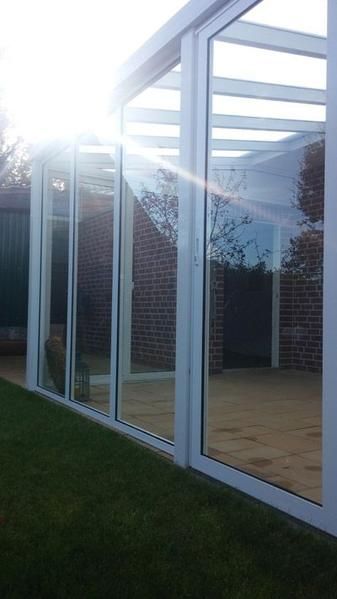 Verglaster Wintergarten mit weißem Rahmen auf grünem Rasen. Helles Sonnenlicht spiegelt sich in den Fenstern. Eine rote Backsteinmauer ist zu sehen.
