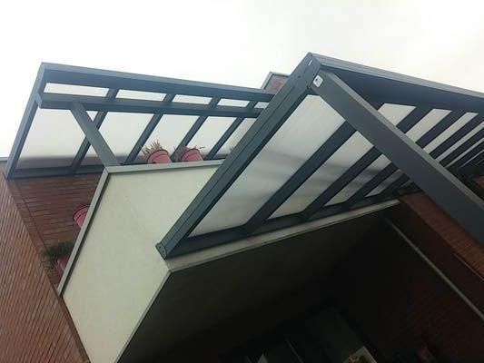 Graue Metallmarkise mit lichtdurchlässigen Dachpaneelen über einem Balkon, der an ein Backsteingebäude angebaut ist.