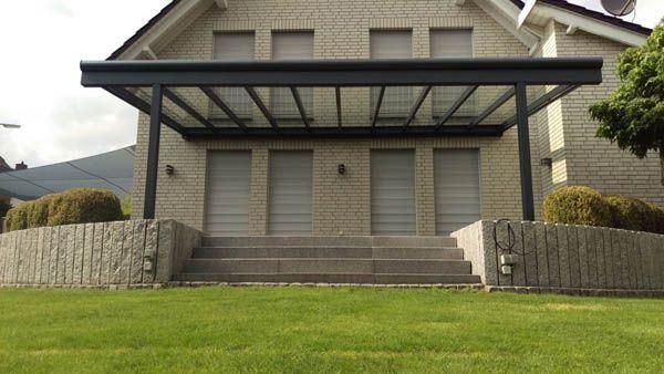 Eine graue Terrassenüberdachung erstreckt sich von einem hellen Backsteingebäude mit drei Türen und einer Treppe, die zu einem Rasen führt.