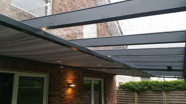 Pergola mit ausfahrbarer Markise über einer Terrasse, Backsteinmauer und Glasfenstern. Grau und Beige.