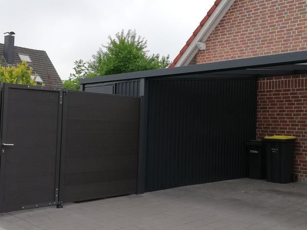 Garage aus schwarzem Wellblech mit angebautem Vordach und dunkelgrauem Tor. Im Hintergrund ein Backsteinhaus.