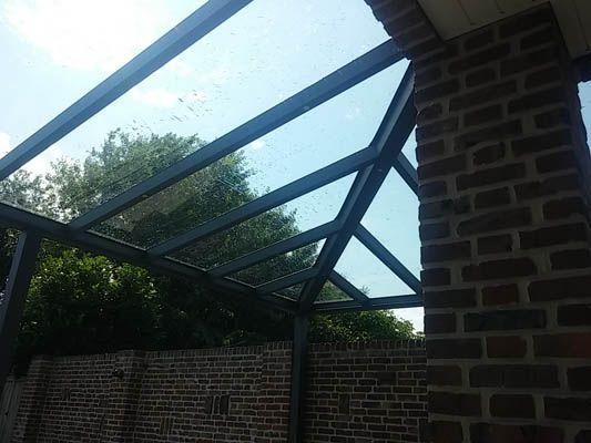 Glasüberdachte Konstruktion mit dunklem Metallrahmen, die an einer Backsteinmauer befestigt ist. Heller Himmel durch das Glas.