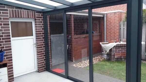 Verglaster Wintergarten mit Backsteinwänden und weißer Tür, der auf einen Rasenhof und ein weiteres Backsteingebäude blickt.