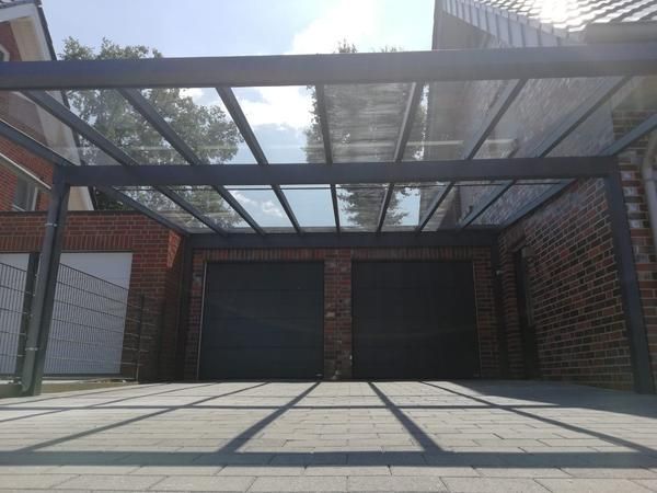 Ein Carport mit Glasdach ist an ein Backsteingebäude mit zwei Garagentoren angebaut; helles Sonnenlicht wirft Schatten.