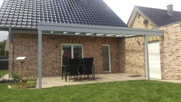 Ein Backsteinhaus mit einem grauen Terrassendach über einer Sitzgruppe. An der Seite befindet sich eine weiße Tür. Davor erstreckt sich grüner Rasen.