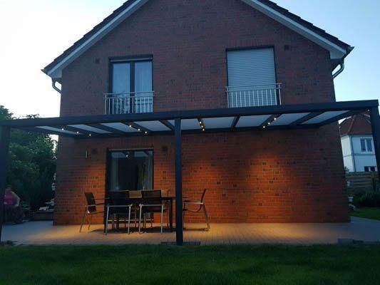 Terrasse mit Tisch und Stühlen unter einer Pergola vor einem Backsteinhaus in der Abenddämmerung, beleuchtet von Deckenleuchten.