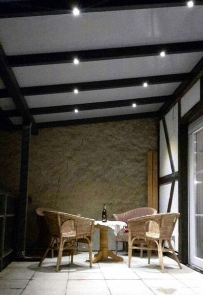 Terrasse mit Korbmöbeln, einem kleinen Tisch und Deckenbeleuchtung. Steinmauer im Hintergrund.