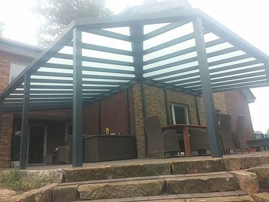 Eine Pergola überspannt eine Terrasse mit Sitzgelegenheiten; im Hintergrund ist ein Gebäude mit Steinfassade zu sehen.