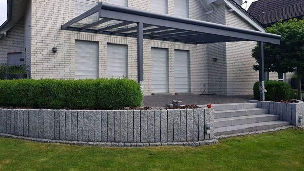 Graue Terrassenüberdachung über einem Türeingang mit einer steinernen Stützmauer und einer Treppe, die dorthin führt.