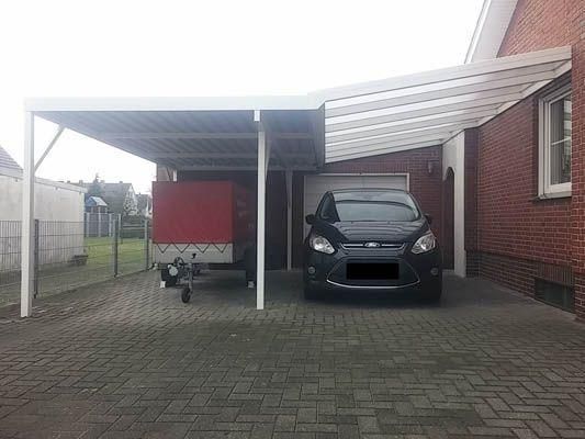 Ein Carport mit einem Auto und einem Anhänger, die auf einer gepflasterten Auffahrt neben einem Backsteinhaus geparkt sind.