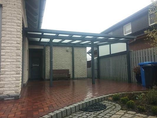 Carport an einem Haus mit gepflasterter Auffahrt. Graues Dach mit transparenten Paneelen. Holzbank.