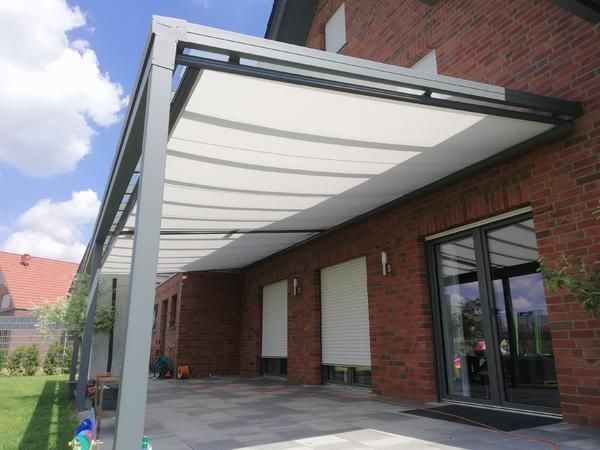 Graue Pergola mit weißem Stoff über einer Backsteinterrasse neben einem Haus.