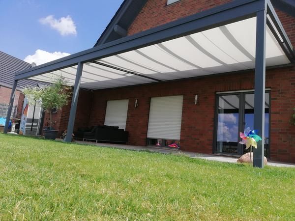 Pergola mit weißem Vordach über einer Ziegelterrasse, vor einem Haus mit grünem Rasen.