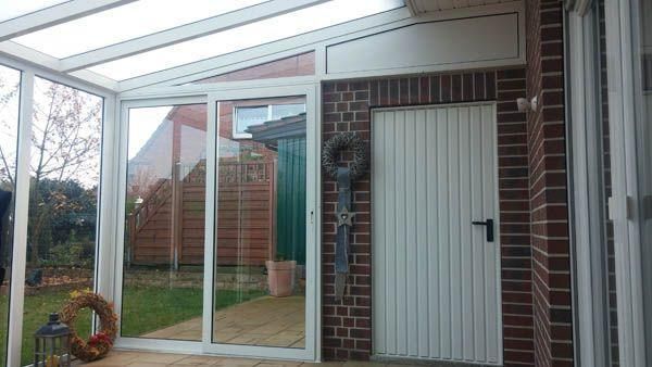 Verglaster Wintergarten mit weißen Zierleisten, Backsteinwand und einer weißen Tür, die nach draußen in den Garten führt.