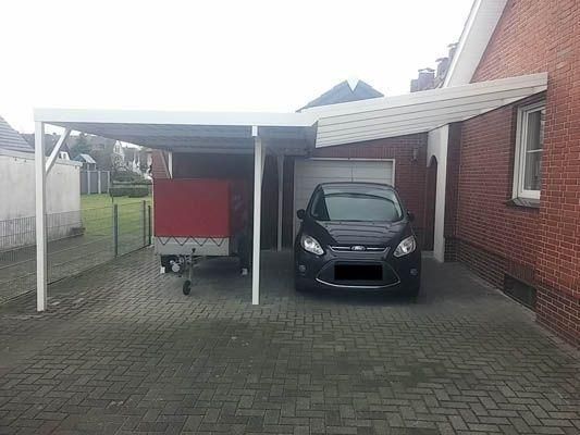 Ein Carport mit einem schwarzen Auto und einem roten Anhänger steht auf einer gepflasterten Auffahrt neben einem Backsteingebäude.